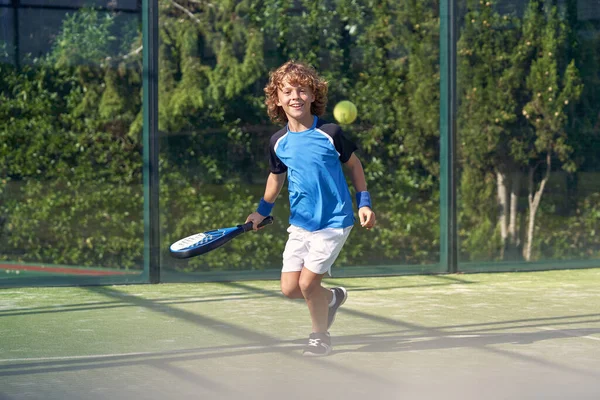 Uzun boylu, kıvırcık saçlı, spor giysili çocuk. Padel raketi ve top oynuyor ve gündüz spor sahasında kameraya bakıyor.