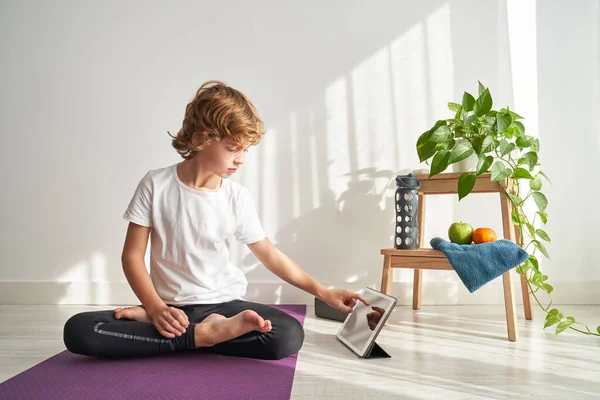 Çıplak ayaklı, kıvırcık saçlı, Lotus yogalı Padmasana asana 'da oturan, bacak bacak bacak üstüne atmış ve ışık odasında sörf tabletinde sörf yapan bir çocuk.