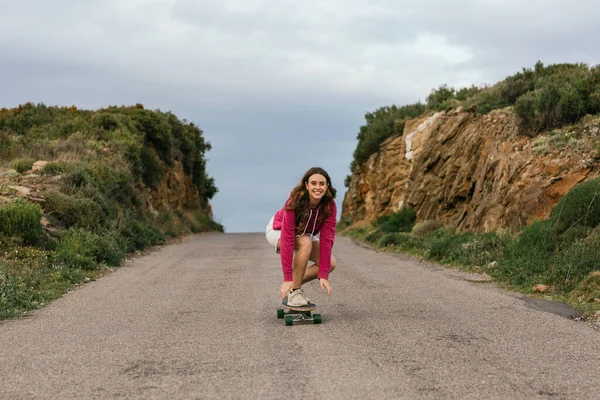 Full body of happy young female in knee pads riding longboard on asphalt road between hills under cloudy sky