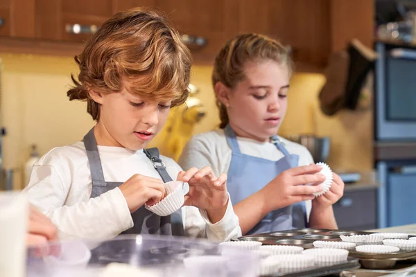 stock image Focused boy and girl preparing baking paper in metal pan for homemade cupcakes while helping crop parent with housework in kitchen