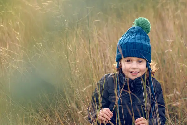 Sıcak şapkalı ve dış giysili pozitif çocuk doğadaki uzun kuru otların arasında dikilirken kameraya gülümseyerek bakıyor.