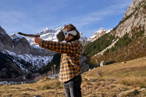 バーチャルリアリティヘッドセットとツリーブランチの男の子の側面のビュー 想像上の銃と高地に立っている間のシミュレーション撮影 — ストック写真