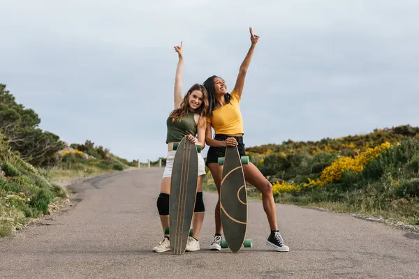 Full body of happy multiethnic girlfriends with outstretch arms standing on road with longboards