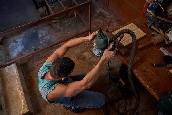 From above of unrecognizable male carpenter crouching at workbench polishing plank using professional sander in workshop