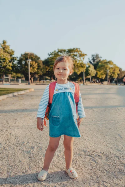 Sırt çantalı, gülümseyen ve parkta kumlu zeminde dururken kameraya bakan sevimli küçük kız.