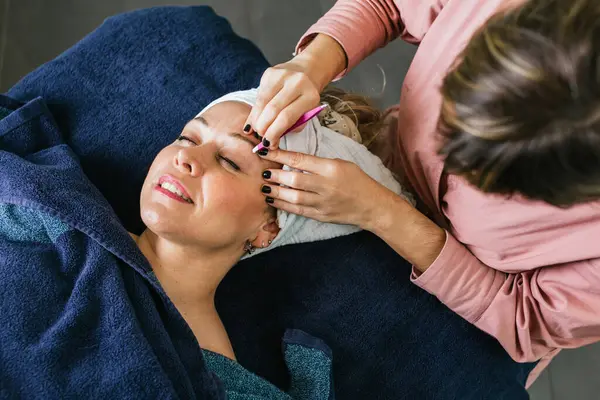 From above of anonymous cosmetologist plucking eyebrows on face of female client lying on table in beauty salon