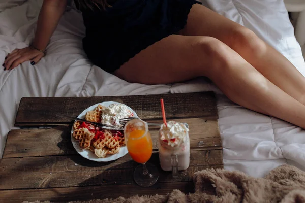 ベッドのトレイで食欲をそそるワッフルやおいしい飲み物を食べる若い女性 — ストック写真