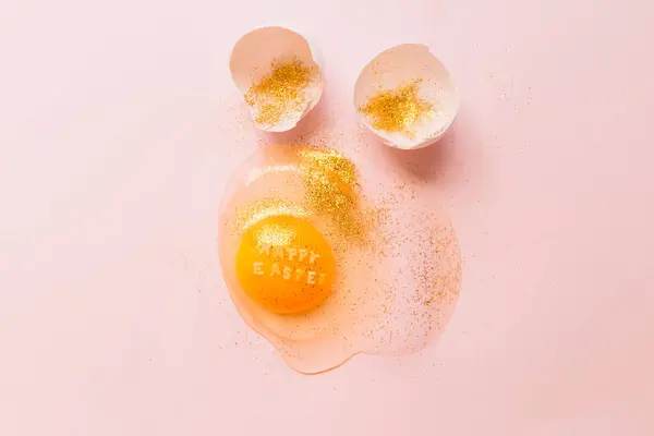 上から生の卵とテキストハッピーイースターに黄色の黄身の輝きとピンクの背景に壊れた卵殻で覆われています — ストック写真