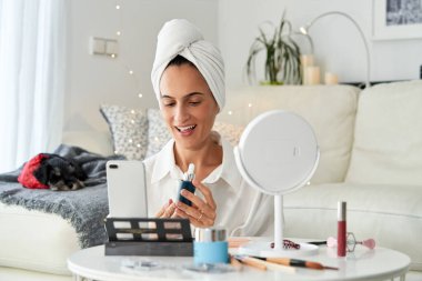 Kafasında havluyla keyifli bir kadın vlogger koltuğun yanında oturuyor ve blog için akıllı telefondan yeni cilt bakım serumu hakkında video çekiyor.