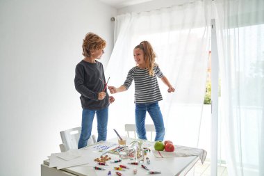 Gülen kız ve erkek kardeş kağıt çarşaflar, akvaryumlar ve güneşli bir sabahta üzerinde akrilik boyalarla beyaz masanın yanında durarak kaynaşıyorlar.