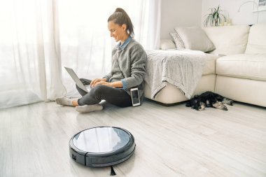 Modern robotik süpürge oturma odasında yerleri temizlerken, uyuyan köpek ve dizüstü bilgisayarın yanında oturan serbest giyinmiş, pozitif kadın çalışanın yan görüntüsü
