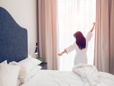 Tanımlanamayan siyah saçlı, bornozlu, pencerenin önünde duran ve sabah vakti otel odasında uyandığında kollarını esneten bir kadının arka görüntüsü.