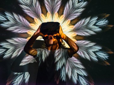 Ağzı açık, VR kulaklık takmış, sanal gerçekliğin tadını çıkarırken parlak neon soyut projektör resminin altında duran komik çocuk.