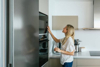 Modern mutfak gereçleriyle mutfakta dikilirken modern siyah mikrodalga fırını açan gündelik kıyafetli bir kadının yan görüntüsü.