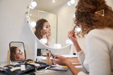 Güzellik stüdyosunda profesyonel fırçayla yüz makyajı yapan genç bir kadının aynasında yansımanın altından.