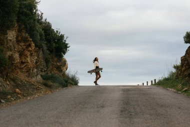 Uzun tahta ve kulaklıklarla ayakta duran, bulutlu gökyüzünün altındaki tepelerin arasından bakan genç, etnik ve neşeli bir kadın.