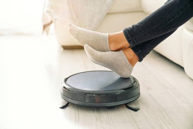 Anonim bir kadının modern bir robot süpürgeye bacak bacak bacak üstüne atması apartmanda ev işi yapması.