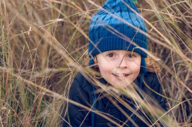 Sıcak giysili pozitif çocuk doğadaki kuru çimenlerde oynarken ve saklanırken kameraya gülümseyerek bakıyor.