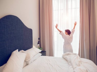 Beyaz bornozlu, tanınmayan bir kadının arkadan görünüşü. Sabahleyin otel odasında uyandıktan sonra pencerenin yanında kollarını gererken.