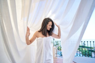 Çekici İspanyol kadın havluya sarılmış otelin terasında uçan tülün altında tatil köyünde yaz günü denize karşı duruyor.
