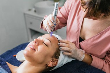 Modern ekipman kullanan, mikro cilt bakımı yapan ve güzellik salonundaki kadın müşterinin yüzünü temizleyen yüksek açılı profesyonel güzellik uzmanı.