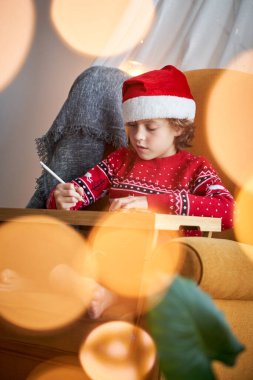 Noel Baba şapkalı küçük çocuk rahat bir koltuğa oturmuş ve masanın üzerine resim çizerken aşağıya bakıyor.