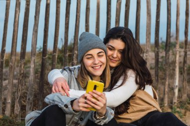 Uzun siyah saçlı, günlük kıyafetlerle kırsal alanda tahta yolda oturan ve akıllı telefon kullanarak en iyi arkadaşına sarılan genç bayan.