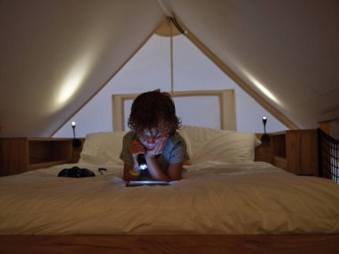 Mansard 'da rahat bir yatakta, evde loş ışık olan ilginç bir kitap okurken gündelik giyinen konsantre çocuk fener takıyor.