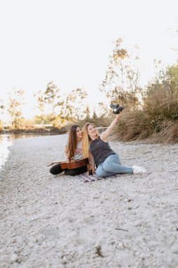 Gitarlı, gülümseyen ve kamerayla selfie çeken genç kadınlar su kenarında kumlu sahilde oturuyorlar.