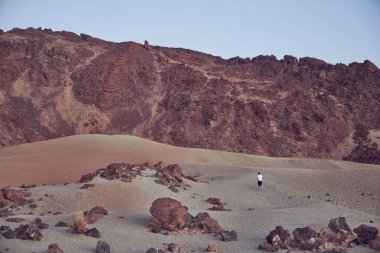 Yaz günü doğa gezisi sırasında kayalık kayalık dağ sırtı yakınlarındaki kumlu arazide mavi gökyüzüne doğru yürüyen uzak bir turist.