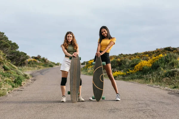 Full body of happy multiethnic girlfriends looking at camera other while walking on road with longboards
