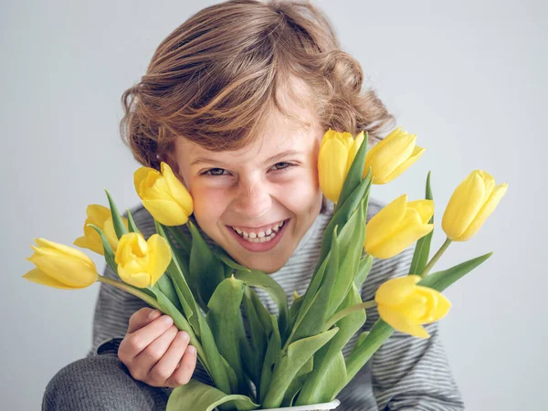 明るい部屋の白い背景に緑の葉が付いている新鮮な黄色のチューリップの花束の近くで笑顔でカメラを見る喜ばしい少年 — ストック写真