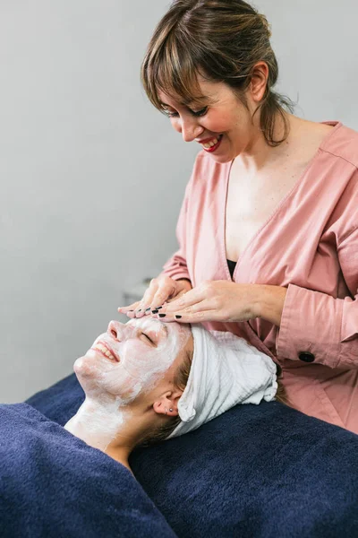 Female cosmetician applying facial cleanser on face of female client during skin care treatment in beauty salon