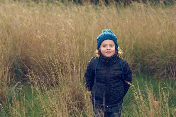 Sıcak giysili ve şapkalı iyimser çocuk, doğadaki kuru çimenlerin yanında durup kameraya gülümseyerek bakıyor.