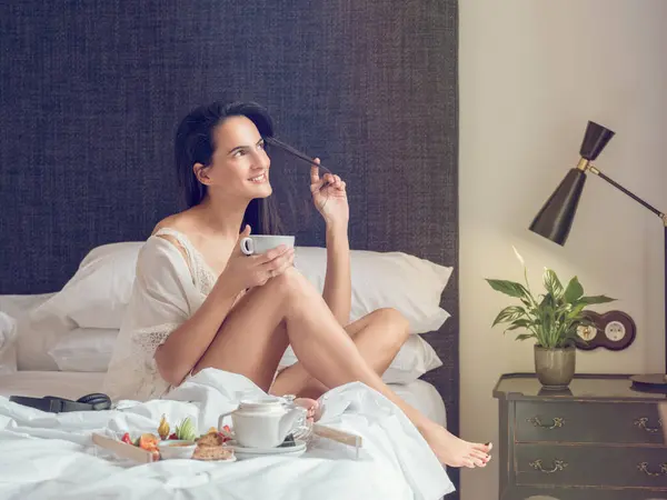 若いセンセーショナルな女性がベッドに座ってナイトウェアを着用し コーヒーと朝食のトレイで朝の寝室を眺めている様子 — ストック写真