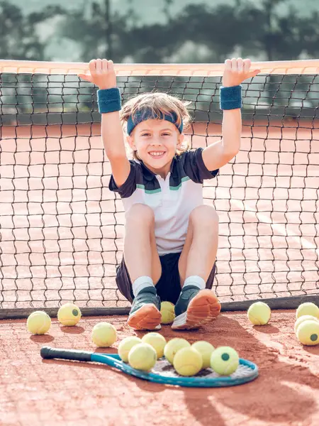 全身上下都是积极的小男孩 金发碧眼 穿着运动鞋 笑着看着镜头 靠在网球场上 坐在网球场上打网球训练 — 图库照片