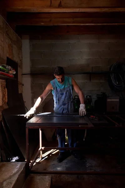 日差しに照らされたみすぼらしいワークショップでプロの楽器を使用しながら金属加工を行う作業服の男性職人の全身 — ストック写真