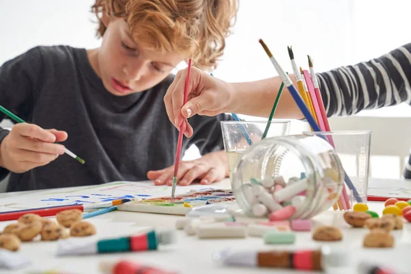 Focused boy painting on paper with colorful paints while sitting at messy table with crop anonymous friend in light room
