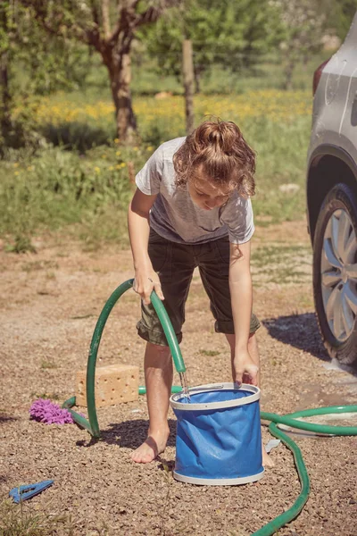 田舎で車を洗っている間 パイプでバケツに水を注ぐ小さな子供の全長 — ストック写真
