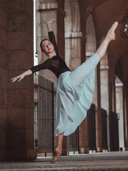 在拱形建筑通道上表演芭蕾舞时 穿着丝裙和尖鞋的全身芭蕾舞演员抬起腿和胳膊的侧视图 — 图库照片