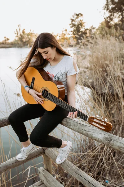 晴れた日に穏やかな湖の近くの木製のフェンスに座っている間 カジュアルな服装でギターを演奏するかなり若い女性 — ストック写真