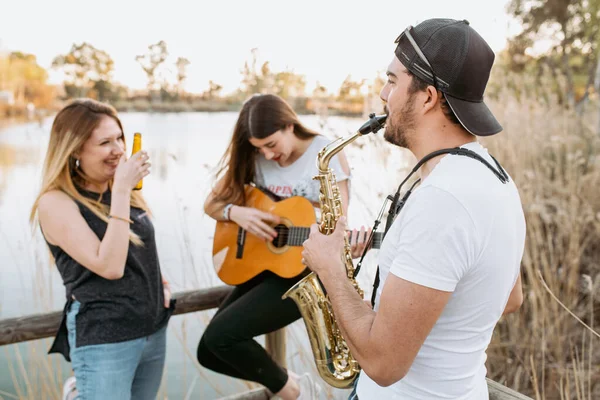 カジュアルな服装の若い女性はビールを飲みながら 湖の近くの晴れた日にギターとサックスを演奏する友人を見て笑っています — ストック写真