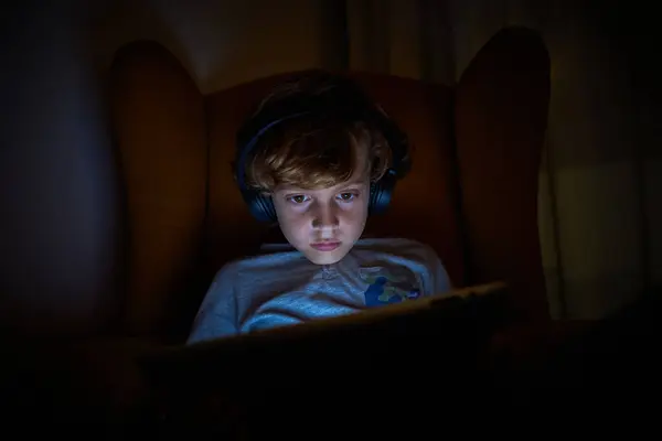 在现代无线耳机里 认真的男孩晚上坐在黑暗的房间里 一边在平板电脑上玩电子游戏 一边坐在椅子上 — 图库照片