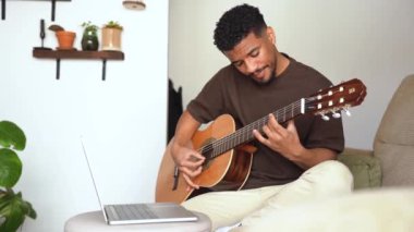 Pozitif Afrikalı Amerikalı erkek müzisyen, gri arka planda rahat bir kanepede akustik gitar çalarken dizüstü bilgisayarda video dersi izliyor.