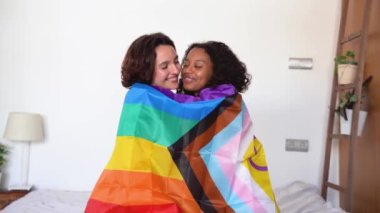 Kameraya bakan, gülümseyen genç lezbiyen çiftin yan görüntüsü. Diğer kucaklaşmalar ve burun uçlarına dokunmalar. Beyaz duvarın etrafında LGBT bayrağını örterken.