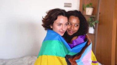 Beyaz duvarın etrafında LGBT bayrağını örterken birbirlerine sarılıp burun uçlarına dokunan gülümseyen çok kültürlü lezbiyen çiftin yan görüntüsü