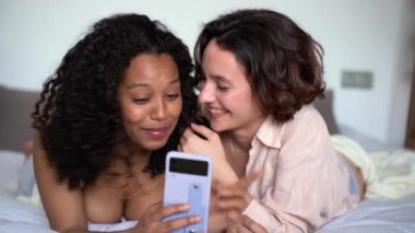 Genç Afro-Amerikan kadın, çeşitli gülümseyen arkadaşlarıyla birlikte beyaz yatakta uzanıp hafta sonunun tadını çıkarırken birlikte cep telefonu ekranına bakıyor.