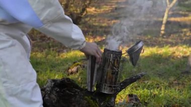 Kırsal kesimde çalışırken arıları sakinleştirmeden önce koruyucu kıyafetli, tanınmayan erkek çiftçinin sigara içmesi