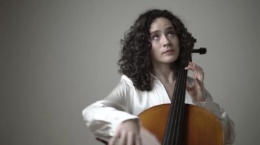 Sandalyeye oturmuş, beyaz duvarlı müzik stüdyosunda keman çalan gündelik kıyafetli genç bir kadın.