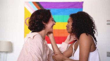 LGBT bayrağına karşı otururken birbirine gülümseyen ve dokunan neşeli, çok ırklı genç bayanların yan görüntüsü